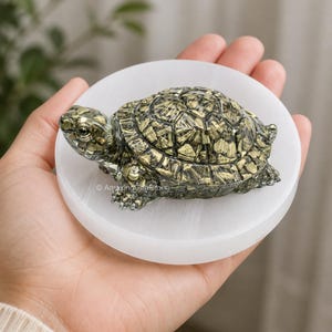 May include: A detailed, metallic-gold turtle figurine rests on a round, white, stone dish. The turtle has a textured shell and detailed features. The dish is held in a person's hand. The text "Amazing Gemstone" is visible on the dish.