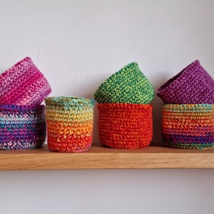 May include: Five crocheted baskets in various colours, including pink, green, orange, and purple, are displayed on a wooden shelf. The baskets are stacked in pairs, with the top basket of each pair slightly tilted forward.