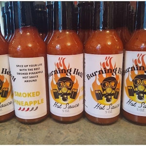May include: A row of six glass bottles of hot sauce labeled "Burning Hell" with a cartoon image of a person wearing a gas mask. The bottles are filled with a red liquid and have white labels with black text. One bottle is labeled "Smoked Pineapple Hot Sauce".