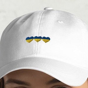 Puede incluir: Gorra de béisbol blanca con tres corazones bordados en los colores de la bandera de Ucrania.