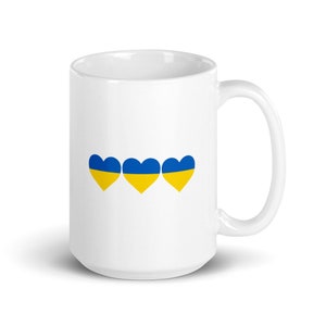 Puede incluir: Taza de cerámica blanca con tres corazones azules y amarillos, que representan la bandera de Ucrania.