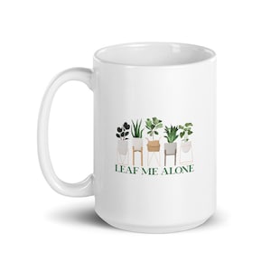 Może przedstawiać: Biały kubek ceramiczny z zielonym nadrukiem przedstawiającym rośliny doniczkowe i tekst "LEAF ME ALONE".