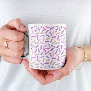 May include: White ceramic mug with a colorful confetti pattern. The confetti is in shades of pink, purple, and gray.