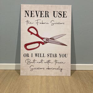 May include: A sign with a humorous warning: "NEVER USE the Fabric Scissors OR I WILL STAB YOU But not with these Scissors obviously..."  The sign features a drawing of a pair of red-handled scissors.