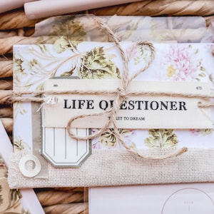 May include: A bundle of stationery with floral patterns and the text "LIFE QUESTIONER". The bundle is tied with twine and includes tags and a burlap strip. The background is a woven basket.