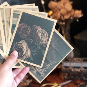 May include: A hand holds a stack of vintage-style postcards with sepia-toned images. The postcards feature floral and architectural designs. The cards have a dark green background and a sepia-toned border. The overall aesthetic is vintage and romantic.