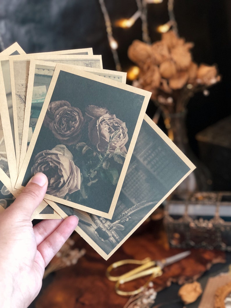 May include: A hand holds a stack of vintage-style postcards with sepia-toned images. The postcards feature floral and architectural designs. The cards have a dark green background and a sepia-toned border. The overall aesthetic is vintage and romantic.