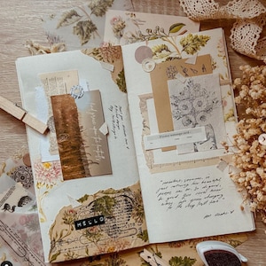May include: Open journal with vintage-style scrapbook elements. Features torn paper, floral patterns, and handwritten notes. Includes a landscape photo, a "HELLO" label, and a "Flower message card-". Wooden clothespins and dried flowers add to the aesthetic.