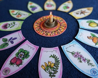 May include: A set of 12 illustrated cards depicting different plants and flowers, arranged in a circle around a lit candle. Each card features a colorful illustration of a plant, along with its name and the month it blooms. The cards are on a blue surface.