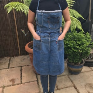 May include: A denim apron with a frilled trim, featuring multiple pockets and a patchwork design. The apron is worn over a black t-shirt.