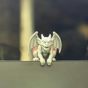 May include: A small, light gray gargoyle figurine with pointed ears, sharp teeth, and spread wings. The creature is depicted in a crouching position with its front claws resting over the edge of a flat, dark surface, showing detailed facial features.