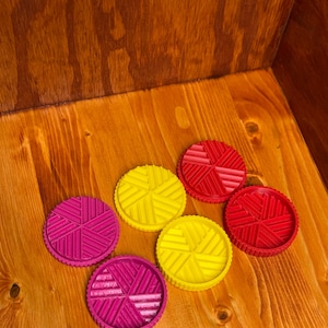 May include: Six colorful, circular game pieces in shades of red, yellow, and magenta are arranged on a wooden surface. Each piece features a geometric design and a textured edge. The pieces are likely for a board game or a similar activity.