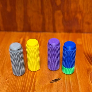 May include: Four colorful, cylindrical objects with textured surfaces. The objects are gray, yellow, purple, and blue with a green base. They are arranged on a wooden surface.
