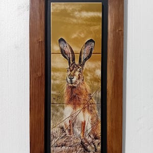 May include: A framed artwork depicting a hare in a field. The hare is rendered in shades of brown and tan, with long ears and a watchful gaze. The artwork is set against a golden backdrop and framed in a dark brown wooden frame.