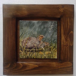 Può includere: Una piastrella incorniciata con un dipinto di due pernici grigie in un campo erboso. La piastrella è circondata da una cornice in legno marrone scuro.
