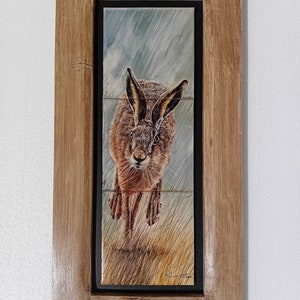 May include: A framed tile artwork featuring a brown and white hare running through a field with a blue and grey sky with rain. The tile is set in a wooden frame.