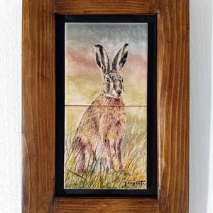May include: A framed tile featuring a watercolour painting of a brown hare sitting in a field of long grass. The background is a soft sunset sky with shades of pink, orange, and blue. The tile is framed in a dark brown wooden frame.