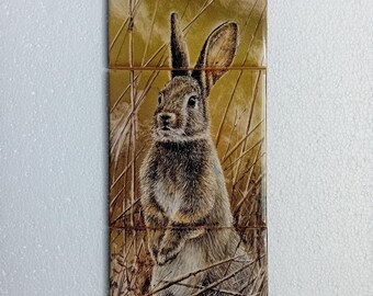 Könnte beinhalten: Ein rechteckiges Fliesenset mit einer detaillierten Illustration eines Kaninchens in einer natürlichen Umgebung. Das Kaninchen hat braunes und graues Fell, lange Ohren und ist von hohem, trockenem Gras umgeben. Der Hintergrund ist ein gedämpftes Olivgrün.