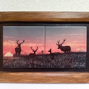 Può includere: Opera d'arte incorniciata con sagome di cervi contro un tramonto. L'immagine è divisa in due piastrelle quadrate, incorniciate in una cornice marrone scuro. Il cielo passa dal rosa al grigio, con il sole all'orizzonte.