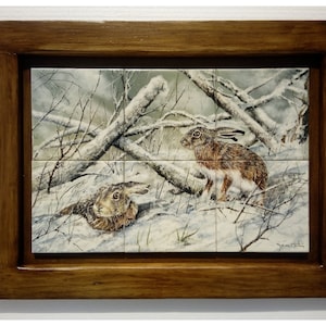 May include: A framed tile artwork featuring two brown hares in a snowy woodland scene. The hares are standing in the snow, surrounded by bare branches and snow-covered ground. The artwork is framed in a dark wood frame.