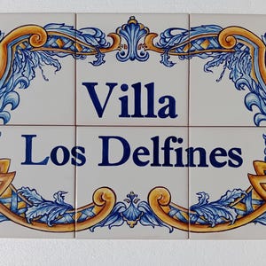 Può includere: Un set di sei piastrelle di ceramica che formano un'insegna. L'insegna recita "Villa Los Delfines" in lettere blu navy. Le piastrelle sono bianche con un bordo decorativo in oro e blu, con disegni floreali e a volute.