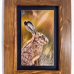 May include: A framed painting of a hare in a field. The artwork depicts the hare with brown and tan fur, long ears, and a detailed eye. The background shows tall grass and a golden sky. The frame is made of stained wood.