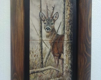 May include: A framed watercolor painting of a deer in a forest setting. The deer is brown with white markings and has antlers. The background is a muted green and brown with trees and brush.
