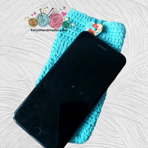 May include: A turquoise crocheted phone case with a floral button. The case is holding a black smartphone.
