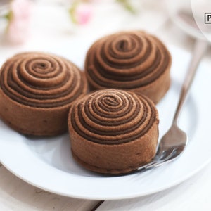 Könnte beinhalten: Drei Schokoladen-Swirl-Kuchen auf einem weißen Teller. Die Kuchen sind in einer Reihe angeordnet, wobei der größte Kuchen hinten und der kleinste Kuchen vorne liegt. Die Kuchen sind alle mit einem spiralförmigen Schokoladenmuster verziert.