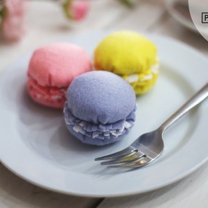 May include: Three felt macarons on a white plate with a silver fork. The macarons are pink, yellow, and purple.