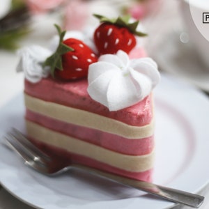 May include: A pink and white felt cake slice with white frosting and two red strawberries on top. The cake is on a white plate with a silver fork.
