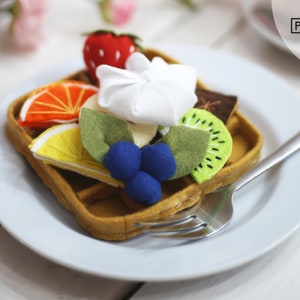 May include: A felt waffle with a variety of fruit toppings, including a strawberry, orange, lemon, kiwi, blueberries, and a dollop of white frosting. The waffle is on a white plate with a silver fork.