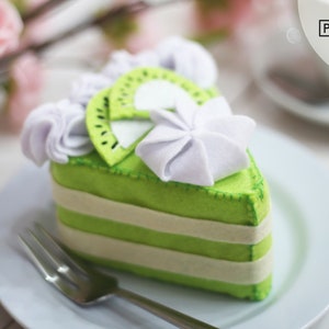 Può includere: Una fetta di torta di feltro verde con glassa bianca e una fetta di kiwi in cima. La torta è su un piatto bianco con una forchetta.