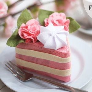 May include: A pink felt cake slice with white frosting and pink felt flowers on top. The cake is on a white plate with a silver fork.