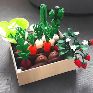 Può includere: Una scatola di legno piena di verdure e fragole di feltro. La scatola contiene lattuga verde, carote, ravanelli, cipolle e una pianta di fragole con fragole rosse.