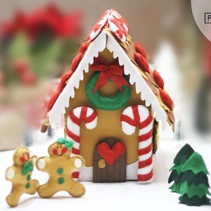 May include: A felt gingerbread house with white icing, red and white candy canes, a green wreath, and a red heart door. Two gingerbread people stand in front of the house, and a green felt Christmas tree is to the right.