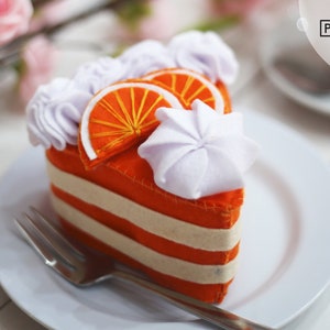 Può includere: Una fetta di torta all'arancia in feltro con glassa bianca e fette d'arancia in cima. La torta è su un piatto bianco con una forchetta d'argento.