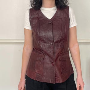 May include: A sleeveless burgundy leather waistcoat featuring a V-neckline, three matching circular buttons down the centre front, and two rectangular patch pockets with flaps positioned at the lower front of the garment.