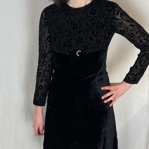 May include: A black velvet dress with long sleeves. The upper bodice features a floral pattern, and the waist is accented with a small, decorative clasp. The dress has a classic, A-line silhouette, perfect for a vintage-inspired look.
