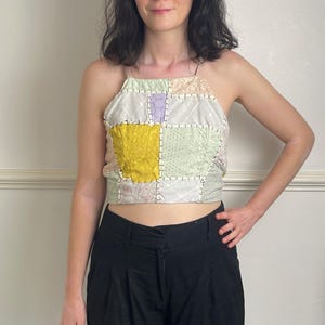 May include: A sleeveless patchwork crop top featuring various fabric squares in shades of yellow, white, light green, and lavender. The pieces are joined together with visible thick white stitching, and the garment has thin spaghetti straps.