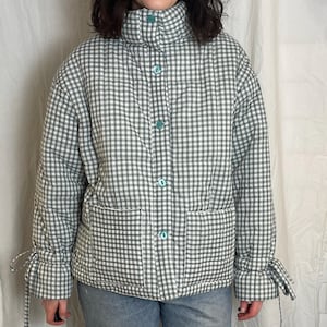Könnte beinhalten: Grün-weiß karierte Jacke mit hohem Kragen und Knopfverschluss. Die Jacke hat zwei Vordertaschen und Bindedetails an den Manschetten. Das Model trägt eine blaue Jeans.
