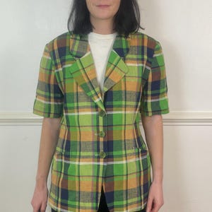 May include: A short-sleeved blazer featuring a vibrant plaid pattern in shades of lime green, mustard yellow, navy blue, and white. The garment includes a notched lapel collar, three front buttons for closure, and two side pockets on the front panel.