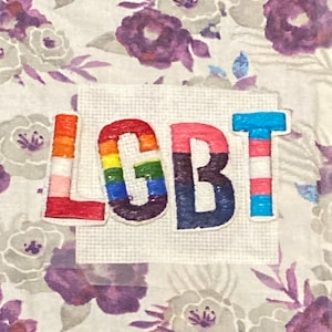 Pode incluir: Patch bordado com as letras "LGBT" nas cores do arco-íris e as cores da bandeira transgênero. O patch está em um fundo branco com um padrão floral em roxo e cinza.