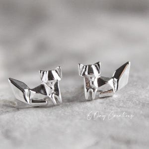 May include: A pair of silver-toned stud earrings shaped like stylized origami foxes. Each earring features geometric facets that form the head, body, and tail of the fox. The small, metallic fox figures are polished to a reflective finish.