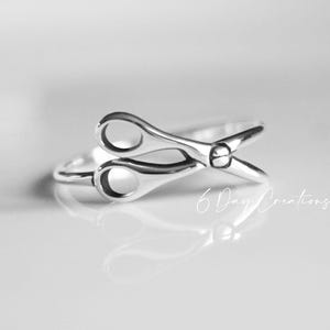 May include: A polished silver ring featuring a design shaped like a pair of open scissors. The metal has a smooth, reflective finish. The text 6 Day Creations is visible in white script in the lower right corner of the image.