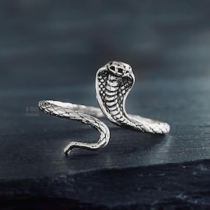 May include: A silver-toned metal ring shaped like a cobra snake. The ring features an open-ended design where the snake's head is raised and the body wraps around to form the band, displaying detailed scale textures across the entire surface.