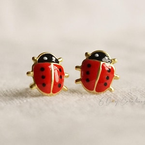May include: A pair of stud earrings shaped like ladybirds with bright red enamel wings, black spots, and a black head section. The edges and legs of the ladybirds are finished in a gold tone. The text 6 Day Creations is visible in the lower right corner.