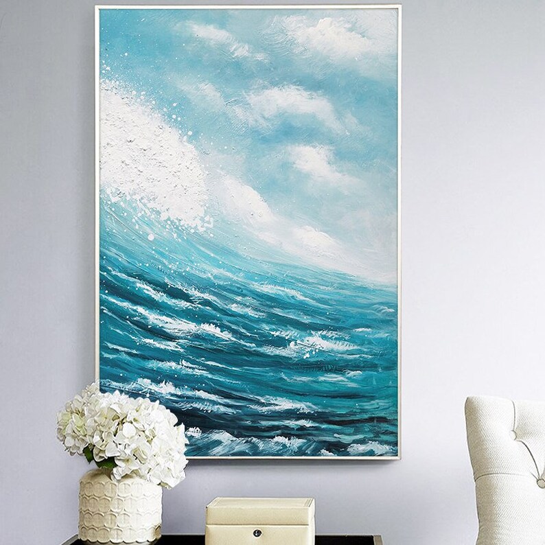 May include: Abstract painting of a blue ocean with white foamy waves crashing against the shore. The painting is framed in a white frame and hangs on a light blue wall.