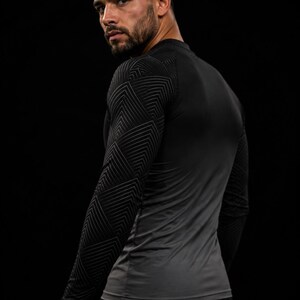 Op de afbeelding: Een compressieshirt met lange mouwen in een zwart en donkergrijs kleurenschema. De mouwen zijn versierd met een geometrisch patroon van dunne, herhalende witte lijnen, terwijl de romp onderaan overgaat in een effen donkergrijze tint.