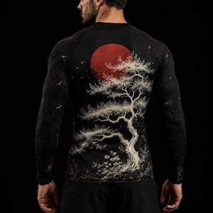Op de afbeelding: Een zwart shirt met lange mouwen en een grafische print op de achterkant. Het ontwerp toont een grote rode zon achter een gedetailleerde witte boom met gestructureerde takken, tegen een donkere achtergrond met kleine witte vogels.